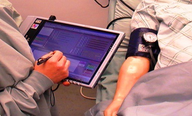 image of healthcare provider's arm and hand using the MTPAT software on a hand held tablet computer with a patient lying next to the provider. you can see the patients right arm with a blood pressure cuff on it.