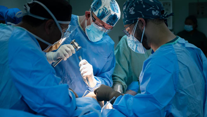 Four surgeons in gowns, caps, and masks perform knee surgery.